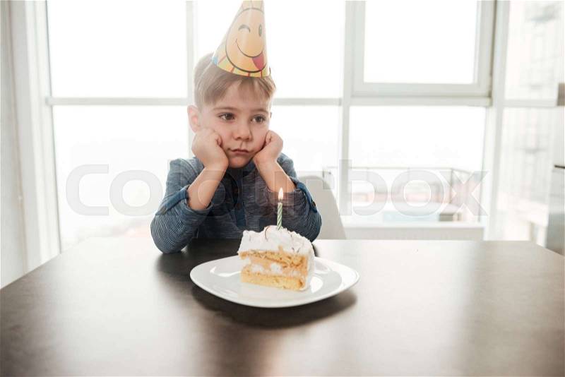 Image of little sad birthday boy sitting in kitchen near cake alone. Look aside, stock photo