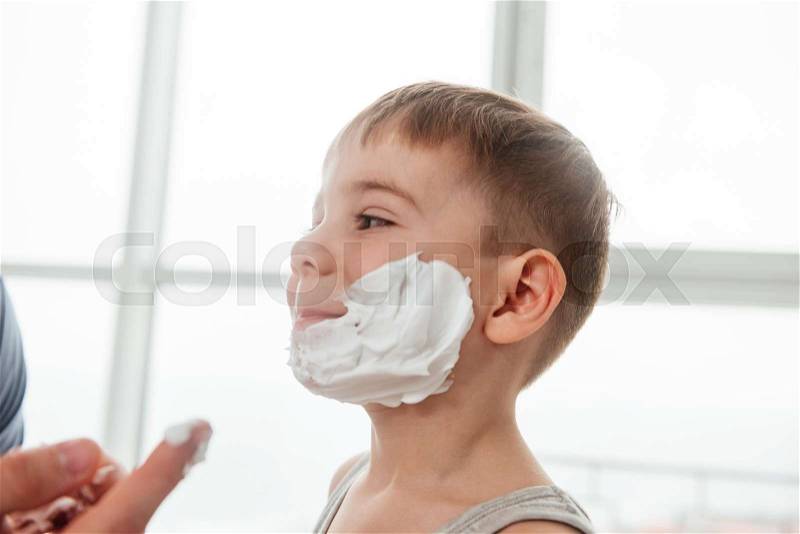Father and son are applying shaving foam on their faces and smiling while shaving in bathroom, stock photo
