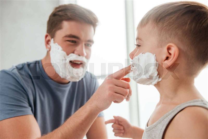Photo of bearded father and son are applying shaving foam on their faces and smiling while shaving in bathroom, stock photo