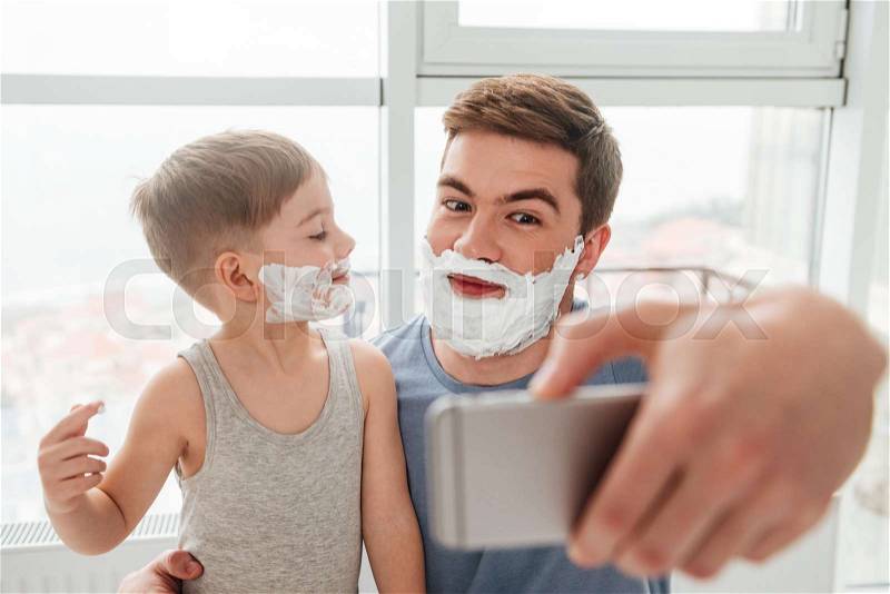 Image of bearded father and son are applying shaving foam on their faces and smiling while taking selfie, stock photo