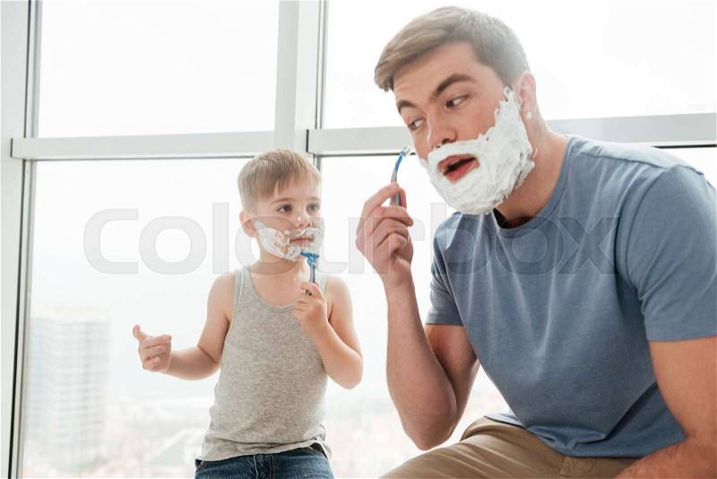 Picture of young father and son are applying shaving foam on their faces and smiling while shaving in bathroom, stock photo