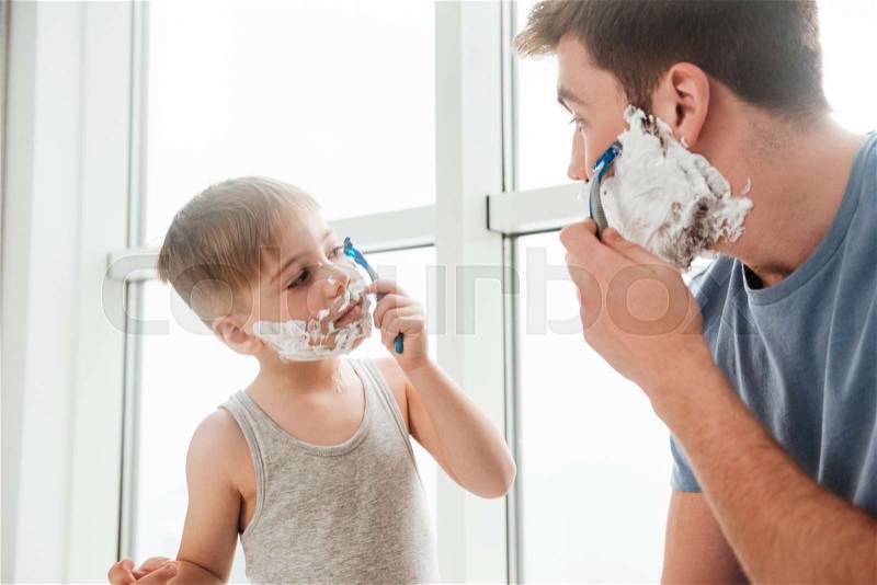 Photo of young happy father and son are applying shaving foam on their faces and smiling while shaving in bathroom, stock photo