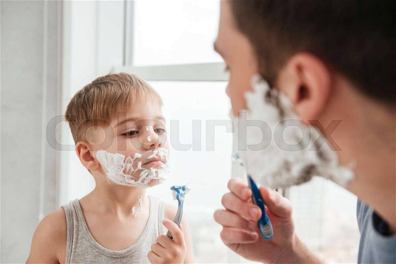 Image of funny father and son are applying shaving foam on their faces and smiling while shaving in bathroom, stock photo