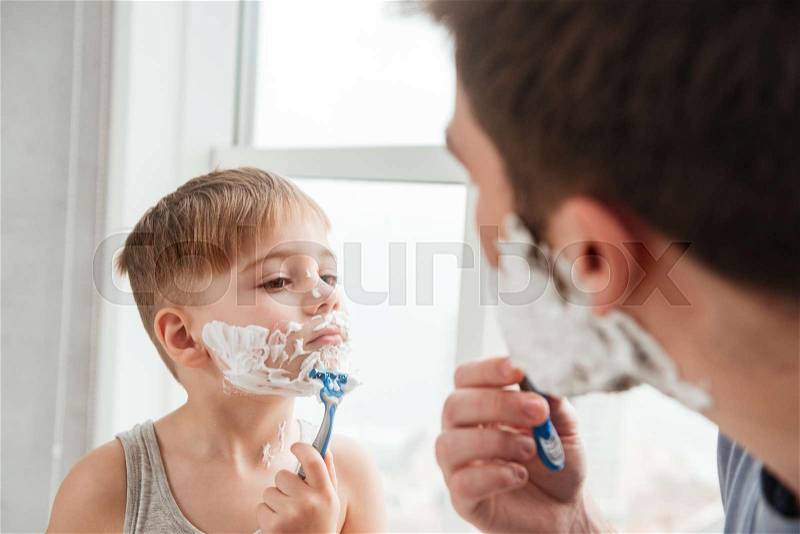 Portrait of funny father and son are applying shaving foam on their faces and smiling while shaving in bathroom, stock photo