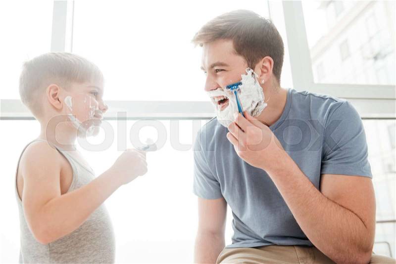 Picture of cute father and son are applying shaving foam on their faces and smiling while shaving in bathroom, stock photo