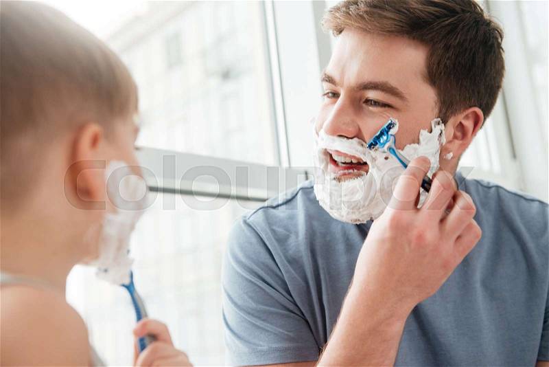 Photo of happy handsome father and son are applying shaving foam on their faces and smiling while shaving in bathroom, stock photo