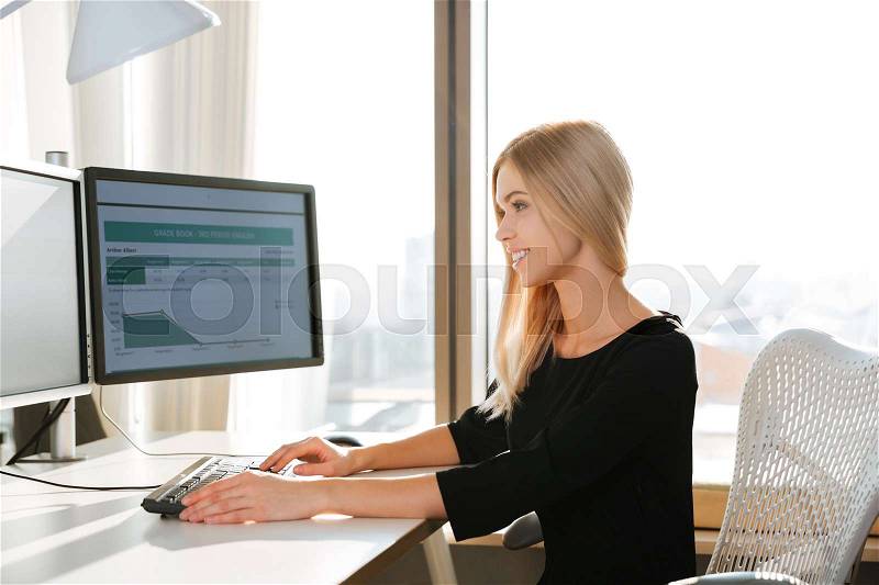 Photo of happy young woman worker using computer in office and typing by keyboard. Look at computer, stock photo