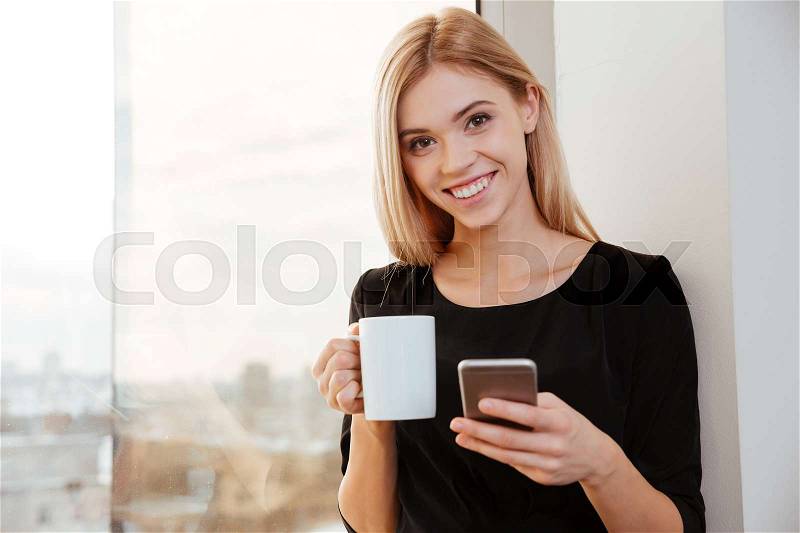 Photo of cheerful lady worker standing in office holding cup of coffee while chatting by her phone. Look at camera, stock photo