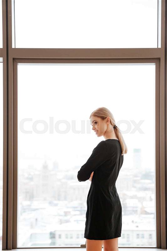 Back view image of young woman worker standing in office near window and looking aside, stock photo