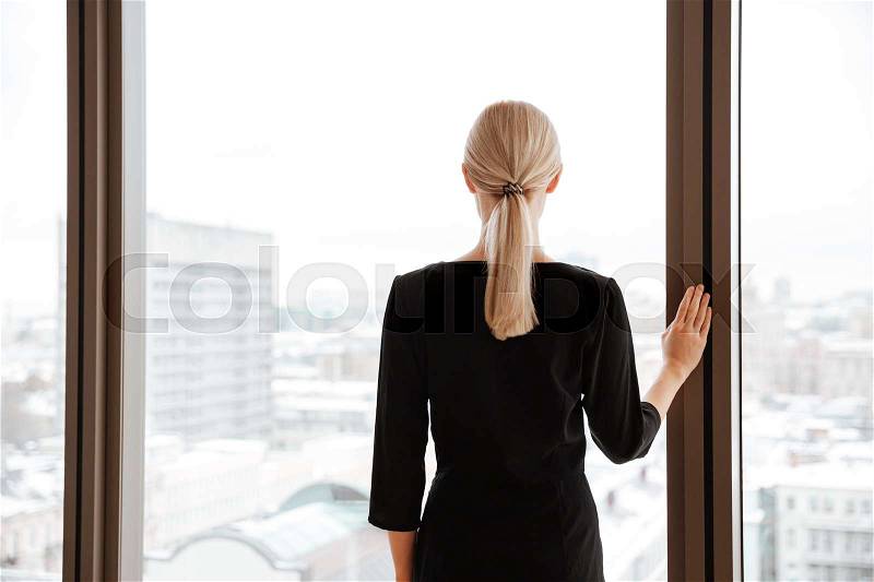 Back view picture of young lady worker standing in office while looking at window, stock photo
