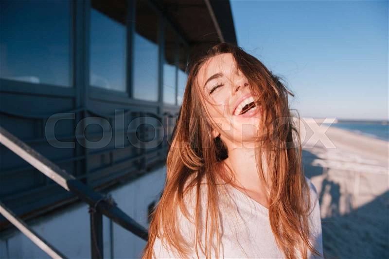 Laughing Woman in sweater standing near the cafe on beach with eye closed and open mouth, stock photo