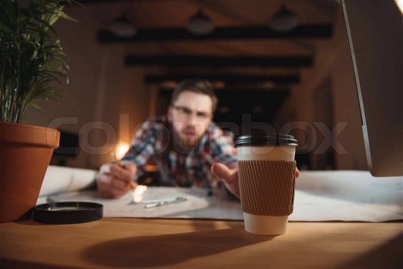 Tired bearded man trying to reach coffee cup while working on desk with graphs, focus on coffee cup, stock photo