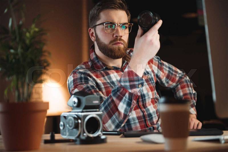 Concentrated young man holding lens while fixing vintage camera at his workplace, stock photo