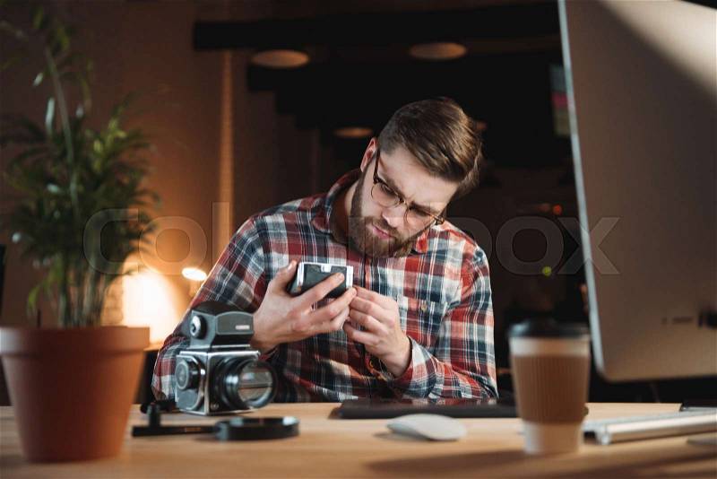 Young bearded man in plaid shirt trying to fix old camera while sitting at his workplace, stock photo