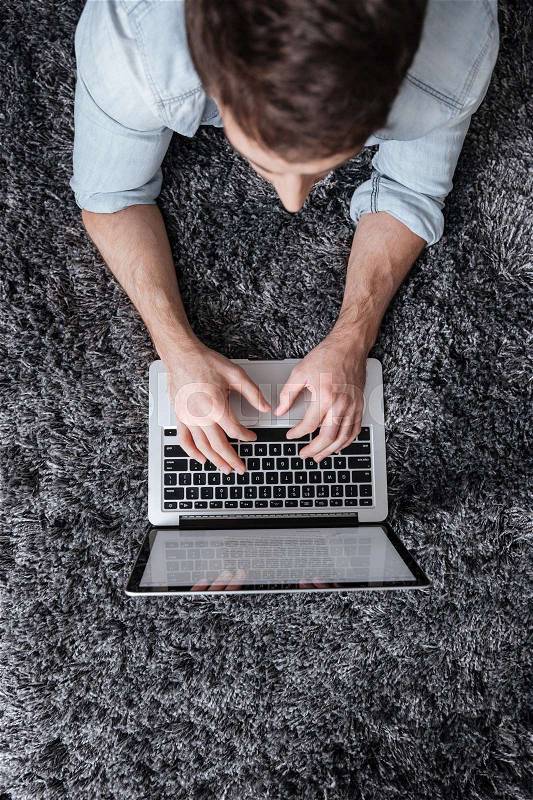 Top view of a young man typing on laptop computer while lying on the carpet at home, stock photo