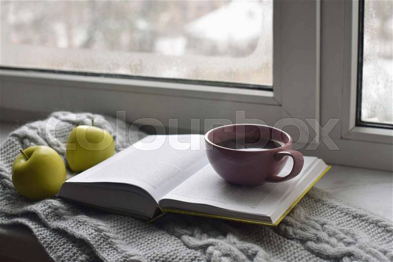 Cozy home still life: cup of hot coffee, green apples and opened book with warm plaid on windowsill against snow landscape outside. Winter holidays and Christmas time concept, free copy space, stock photo