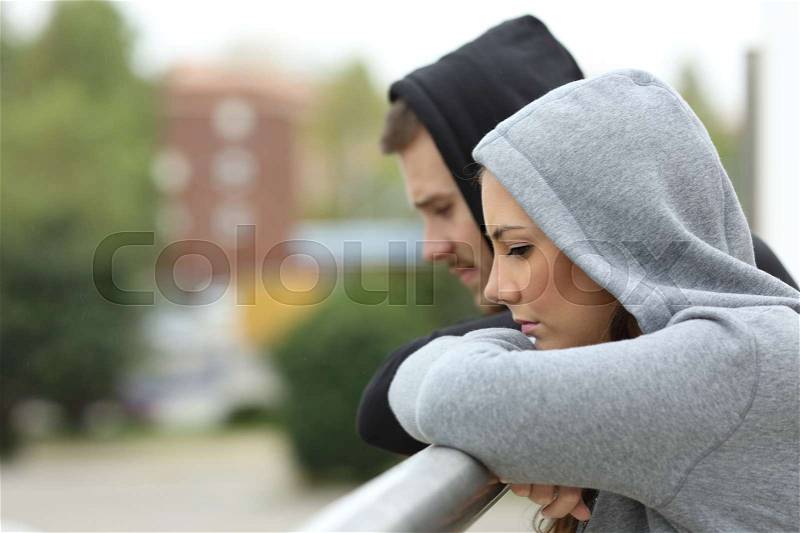 Side view of a sad couple of teens looking down after break up in a balcony of a house with an urban background, stock photo