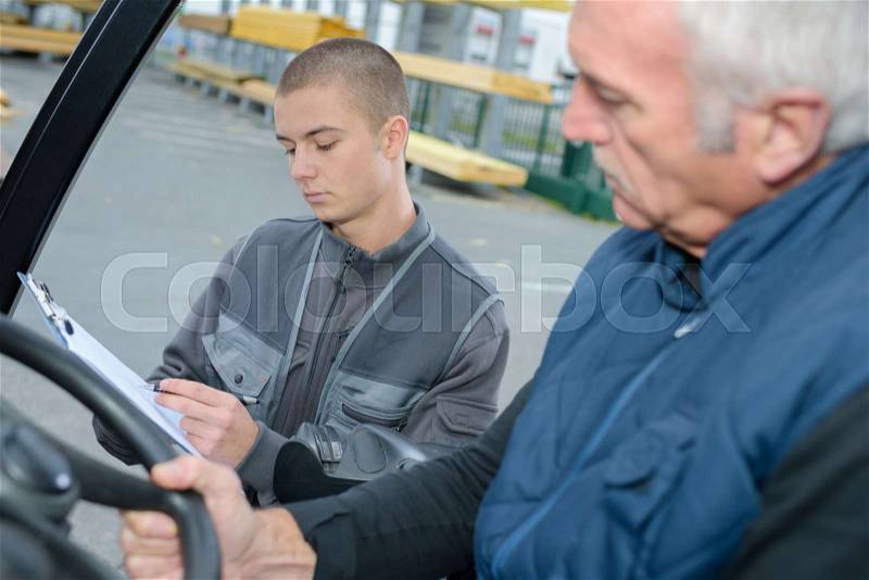 Young man with clipboard next to senior man driving forklift, stock photo