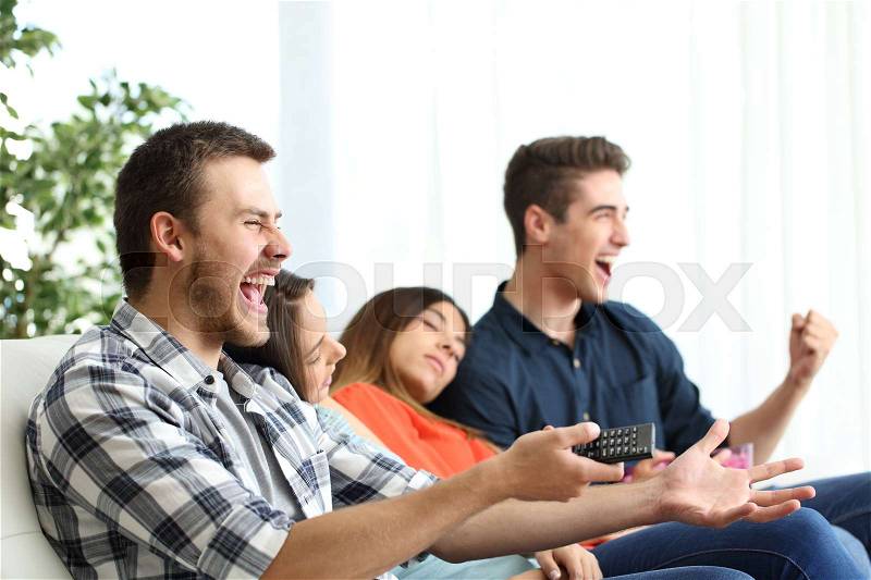 Excited men watching tv and bored girlfriends sitting on a couch in the living room at home, stock photo