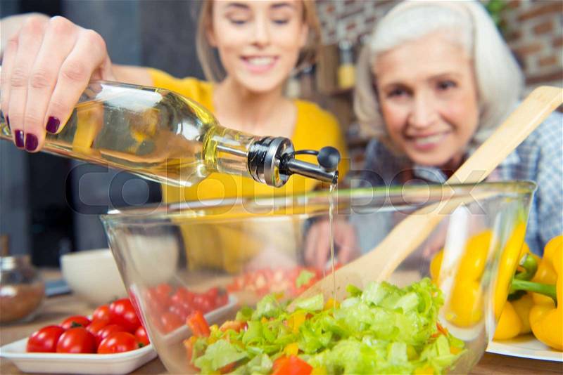 Granddaughter and grandmother cooking together and pouring oil into vegetable salad, stock photo