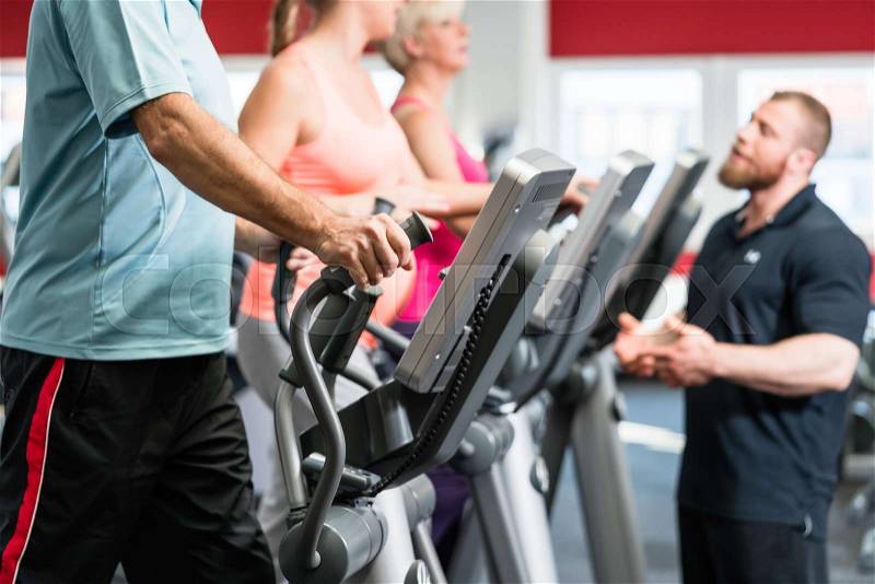 Seniors training on cross trainer with personal trainer at the gym, stock photo