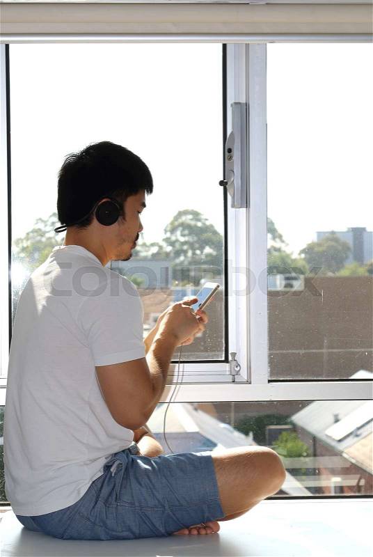 Young Asian man sits by a window which looks over a neighborhood listening to music using headphones, stock photo