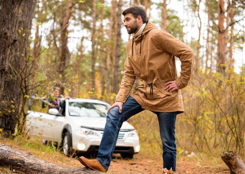 Young bearded man standing on the big dry log on forest road with blurred white car on background, stock photo