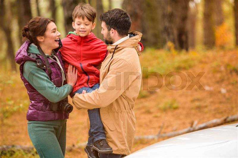 Happy young family standing embracing near white car in autumn forest, stock photo