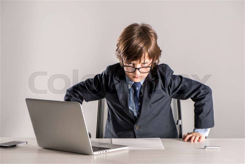Schoolchild in business suit standing over desk and looking angry at camera, stock photo