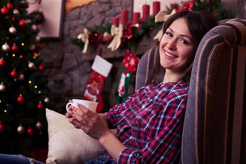 Close-up of overjoyed youth relaxing in an armchair against decorated fireplace. Nice Christmas tree near the fireplace. Festive illumination. Christmas scene with tree in background, stock photo