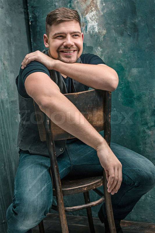 Portrait of smiling happy man sitting on the wooden chair on gray studio background, stock photo