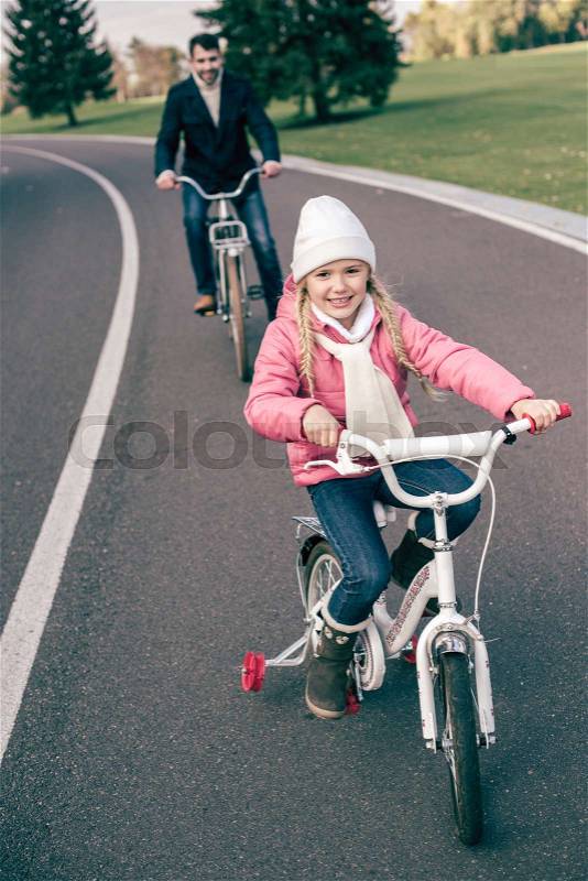 Cute smiling little girl cycling with father on road in autumn park, stock photo