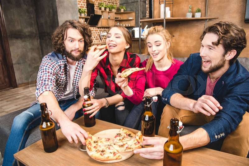 Smiling friends drinking beer and eating pizza while sitting on sofa , stock photo