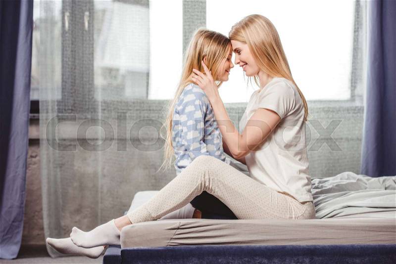 Happy mother and daughter in pajamas sitting head to head on bed , stock photo