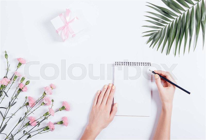 Female hand writing in a notebook at the desk, top view. On the white table lay flowers and a box with a gift and ribbon, stock photo