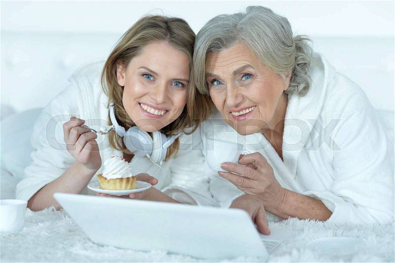 Portrait of senior woman with her adult daughter eat tasty cake, dring tea and watching something on laptop, stock photo