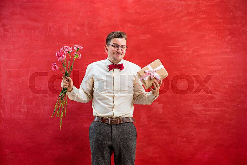 Young disappointed funny man with flowers and gift on red studio background, stock photo