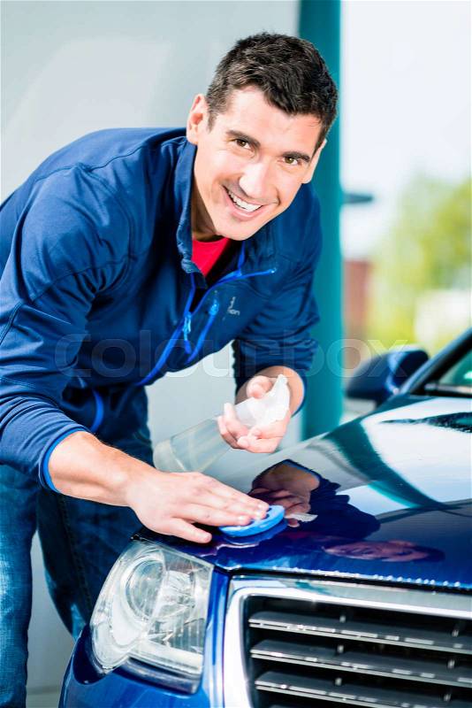 Happy young man looking at camera while waxing a blue car outdoors at car wash, stock photo