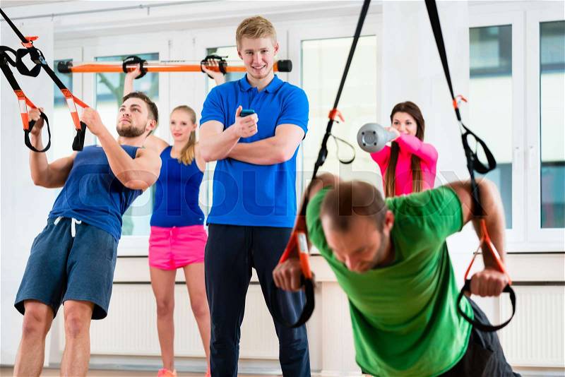 Group of men and women in gym doing workout at rings supervised by physio , stock photo