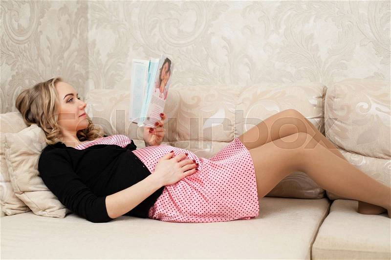 Young beautiful pregnant woman is lying on the back on the sofa and reading a magazine,one hand embracing the belly, stock photo