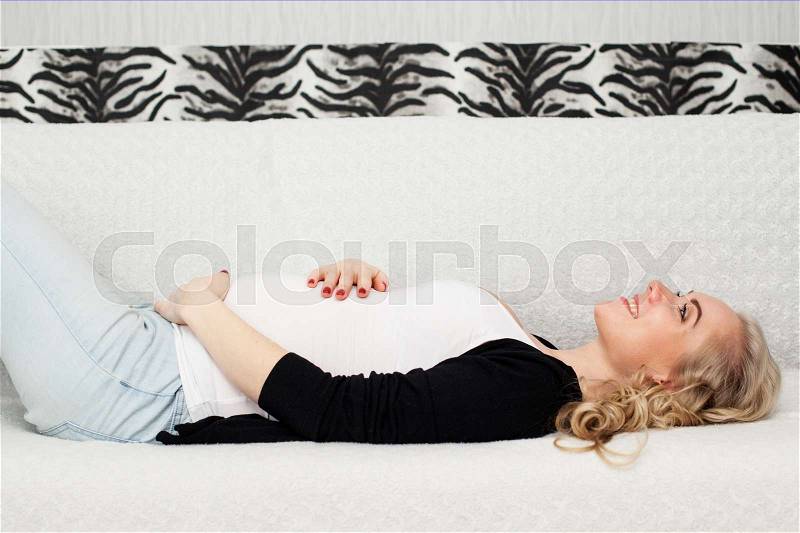 Young beautiful pregnant woman is lying on the back an the sofa and smiling, hands embracing the belly, stock photo