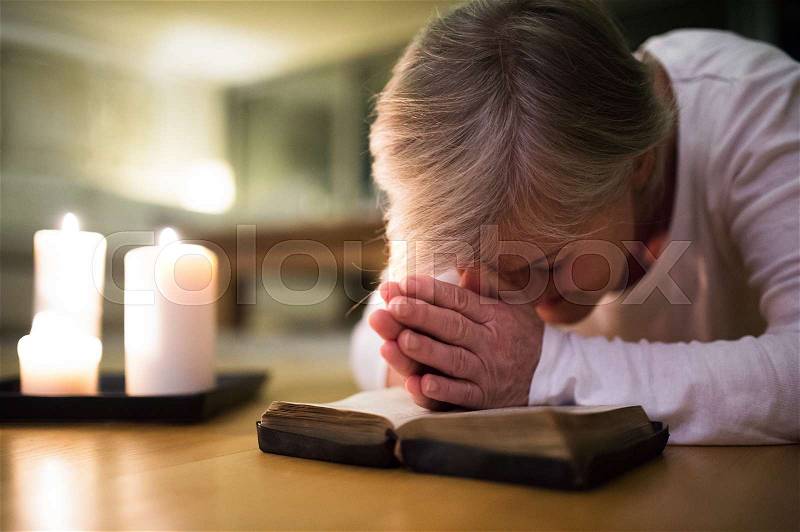 Senior woman kneeling on the floor praying with hands clasped together on her Bible. Burning candles next to her. Close up, stock photo