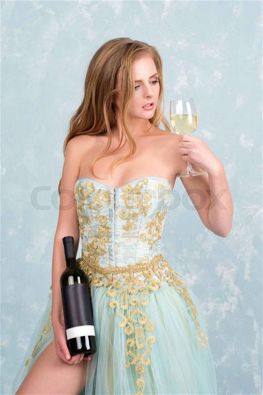 Beautiful sensual blonde woman in gorgeous long dress holding glass of white wine and bottle. Young girl celebrating. Proposing product, advertisement, stock photo