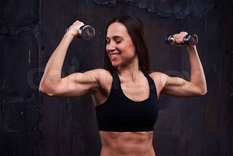 Side view shot of smiling good-looking powerful woman performing sport exercise with double dumbbells. Fitness model accomplishing an intense biceps work out, stock photo