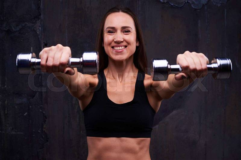Close-up of sporty sportswoman flexing two hands isolated over black background. Trained sportswoman holding two dumbbells on straight hands isolated over black background , stock photo