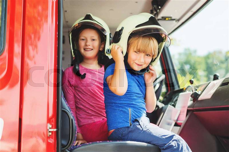 Two cute kids playing in fire truck, pretending to be firefighters, open doors day at fire station. Future profession for children. Educational program for schoolkids, stock photo
