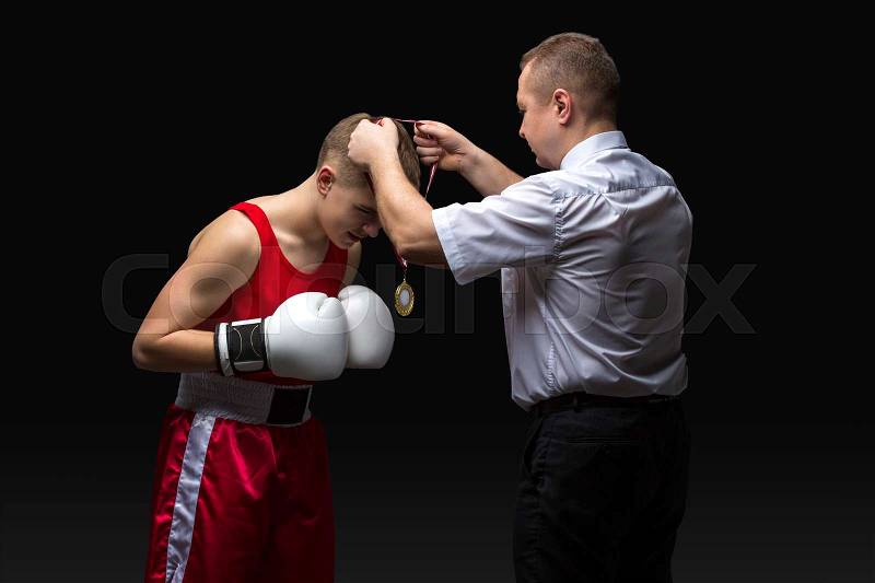 Boxing referee gives medal to young teen boxer in red form and white gloves. Winner. Studio shot on black background. Copy space, stock photo