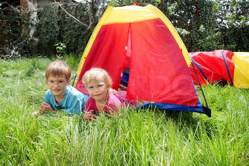 Young brother and sister lying near colorful tent on green grass on summer day, stock photo