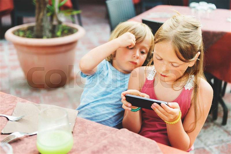 Two kids playing electronic games on the phone while waiting food orders in restaurant, stock photo