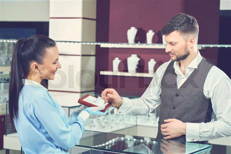Female seller in jewelry store showing engagement ring for buyer. Young brunet man with beard choosing present for his girlfriend, touching for golden ring with blue stone and want buying it, stock photo